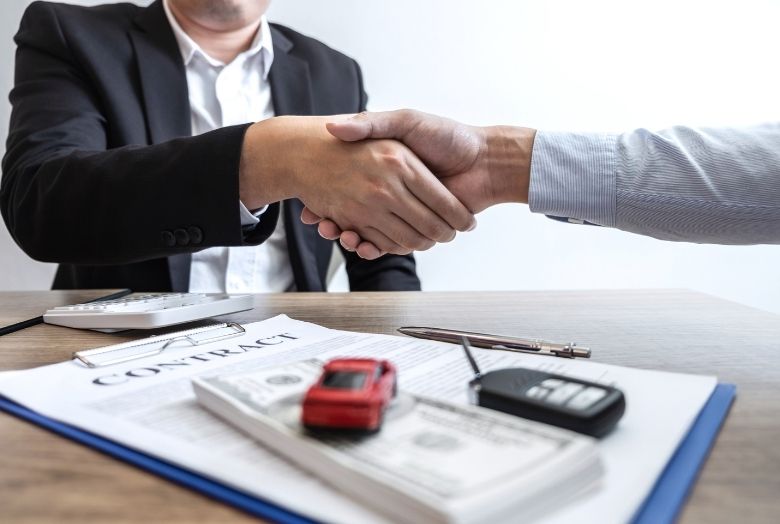 Duas pessoas apertando as mãos fechando negócio em cima de uma mesa com documentos, dinheiro, a chave de um carro e um carrinho em miniatura.